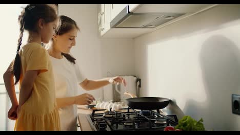 A-brunette-woman-in-a-white-T-shirt-together-with-her-little-daughter-in-a-yellow-dress-tells-her-how-to-properly-use-the-stove-and-cook-food-in-the-kitchen-in-a-modern-apartment