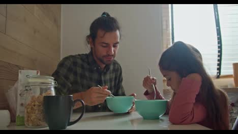 Brunette-man-in-a-green-checkered-shirt,-the-single-father-has-breakfast-with-his-little-daughter,-a-brunette-girl-in-a-pink-dress-at-the-table-in-the-morning-in-a-modern-apartment