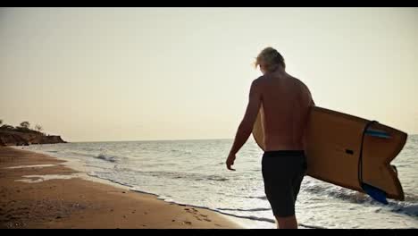 Shooting-on-the-site,-a-blond-man-with-a-bare-torso-in-black-shorts-carries-a-yellow-surfboard-and-walks-along-the-orange-sandy-seashore-in-the-morning-at-Sunrise