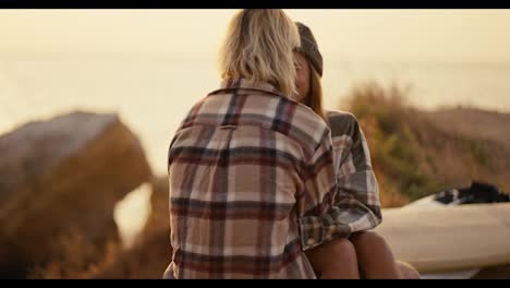 A-blonde-girl-in-a-black-hat-and-a-checkered-shirt-sits-on-the-lap-of-her-blonde-boyfriend-in-a-checkered-shirt-on-the-seashore-Sunny-morning-at-sunrise