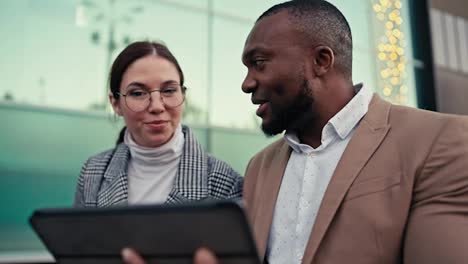 A-confident-man-with-Black-skin-and-a-beard-a-businessman-with-a-short-haircut-in-a-brown-suit-communicates-with-his-colleague-a-brunette-girl-in-round-glasses-and-a-gray-jacket-during-their-meeting-and-viewing-plans-on-a-tablet-near-the-building