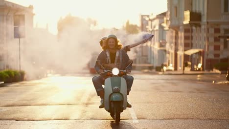 A-happy-guy-with-long-curly-hair-in-a-denim-jacket-rides-on-his-green-moped-with-a-girl-in-a-white-moped-helmet-who-is-holding-a-blue-fire-in-her-hands-that-leaves-behind-a-gray-haze-along-the-summer-morning-street