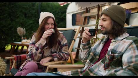 A-happy-brunette-girl-in-a-white-cap-and-a-checkered-shirt-drinks-tea-with-her-brunette-boyfriend-in-a-green-checkered-shirt-sitting-on-special-chairs-during-a-picnic-near-her-trailer-in-the-forest-in-the-summer