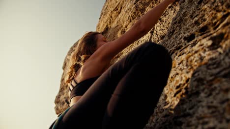 A-blonde-girl-in-a-black-top-and-a-special-harness-on-her-belt-climbs-onto-a-rock-and-then-looks-back-and-down.-Rock-girl-climbing-up-a-rock-with-sunny-summer