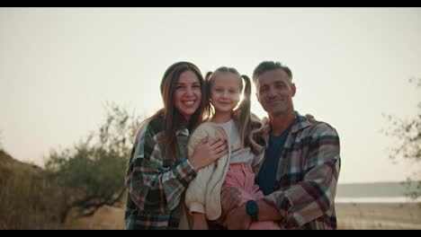 Portrait-of-a-happy-family,-a-little-blonde-girl-sits-in-the-arms-of-her-dad,-a-brunette-man-with-a-little-gray-hair-in-a-brown-checkered-shirt,-next-to-her-is-her-mother,-a-brunette-girl-in-a-green-checkered-shirt.-Family-on-a-hike-and-on-a-picnic-outside-the-city