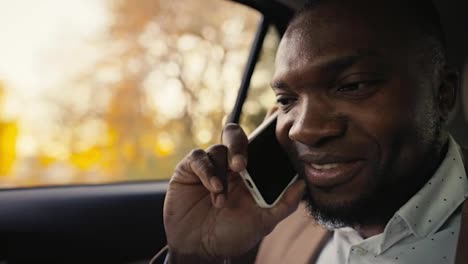 Close-up-of-a-happy-man-Businessman-with-Black-skin-color-in-a-brown-jacket-communicates-on-a-smartphone-while-riding-in-the-back-seat-of-a-modern-car-outside-the-city-during-his-business-trip