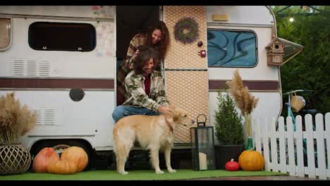 Happy-brunette-guy-in-a-Green-checkered-shirt-pets-his-big-cream-colored-dog-and-a-brunette-girl-touches-his-hair-Near-his-trailer-during-a-picnic-at-a-camp-outside-the-city-in-the-summer