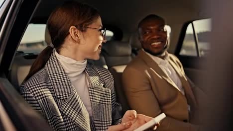 A-happy-brunette-girl-in-round-glasses-rejoices-and-has-fun-while-communicating-with-her-colleague,-a-man-with-Black-skin-in-a-brown-suit-during-his-business-trip-in-a-modern-car