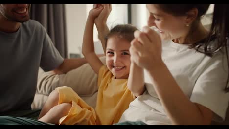 Close-up-shot-of-a-brunette-mom-in-a-white-t-shirt-playing-with-her-daughter-in-a-yellow-dress-while-sitting-on-a-light-brown-sofa-with-her-dad-in-a-modern-studio-room