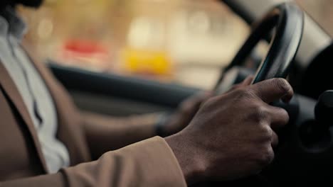 Close-up-shot-of-a-man-Businessman-with-Black-skin-color-in-a-brown-jacket-holds-with-his-two-hands-the-steering-wheel-of-a-modern-car-while-he-is-driving-in-a-car-in-a-modern-salon-during-a-business-trip-in-the-city