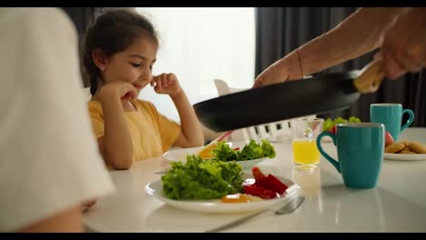 A-man-prepared-a-morning-breakfast-and-puts-scrambled-eggs-on-plates-for-his-little-daughter,-a-brunette-girl-in-a-yellow-dress,-and-for-his-wife,-a-girl-in-a-white-T-shirt,-during-the-morning-breakfast-with-salad-at-the-table-in-the-kitchen