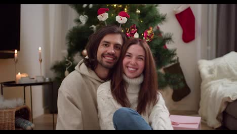 Retrato-De-Una-Pareja-Feliz,-Un-Chico-Moreno-Está-Sentado-Con-Su-Novia-Con-Un-Suéter-Blanco-Y-Adornos-De-Año-Nuevo-Cerca-De-Un-árbol-De-Año-Nuevo-Decorado-Con-Juguetes-De-Año-Nuevo-En-Una-Acogedora-Habitación-En-Invierno.