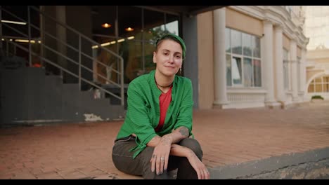 Portrait-of-a-lesbian-girl-with-short-green-hair,-tattoos,-in-a-Green-shirt-and-black-pants-who-sits-on-a-small-step-on-the-street-near-the-building