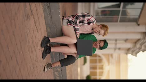 Vertical-video:-two-lesbian-girls-sit-on-the-steps-in-the-city-and-look-at-a-gray-monitor-screen-during-their-date.-A-blonde-girl-with-a-short-haircut-in-a-checkered-pink-shirt,-together-with-her-girlfriend-with-a-short-green-haircut-in-a-green-shirt,-are-talking-and-watching-something-on-the-monito