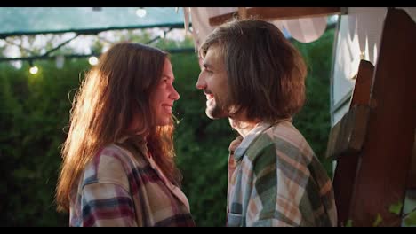 Happy-brunette-girl-together-with-her-brunette-boyfriend-in-plaid-shirts-stand-next-to-each-other-close-and-look-at-each-other-Near-their-trailer-during-a-picnic-in-a-camp-outside-the-city-in-the-summer