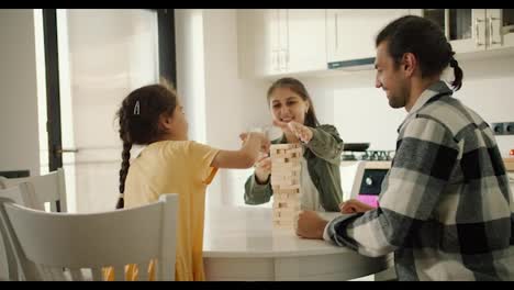 Rear-view-of-a-little-brunette-girl-in-a-yellow-dress-playing-the-board-game-Jenga-with-her-parents-on-the-table-in-a-white-modern-kitchen