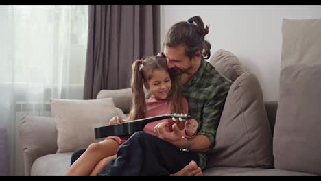 Una-Niña-Morena-Con-Un-Vestido-Rosa-Se-Sienta-En-El-Regazo-De-Su-Padre-Moreno-Con-Una-Camisa-A-Cuadros-Verde-Y-Afina-El-Ukelele-Para-Tocar-Un-Instrumento-Musical-Mientras-Está-Sentada-En-Un-Sofá-Gris-En-Casa-En-Un-Apartamento-Moderno.