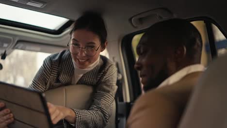 A-happy-and-confident-brunette-girl-in-round-glasses-in-a-suit-communicates-with-a-Black-skinned-businessman-in-a-brown-jacket-and-presents-her-plans-using-a-tablet-during-her-car-ride-and-business-trip