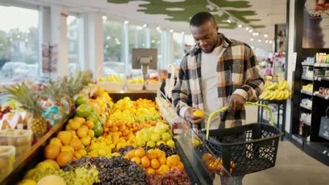 A-guy-with-Black-skin-in-a-checkered-shirt-chooses-citrus-fruits-and-berries-on-the-wide-counter-of-a-modern-supermarket-and-goes-on-with-his-shopping-business