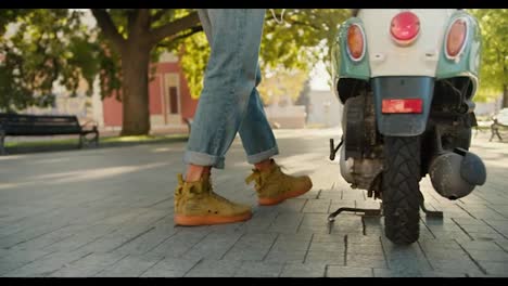 Shooting-close-up,-a-guy-in-denim-shorts-walks-to-his-green-moped,-sits-on-it-and-drives-away-along-the-street-with-trees-and-a-lawn-in-the-summer