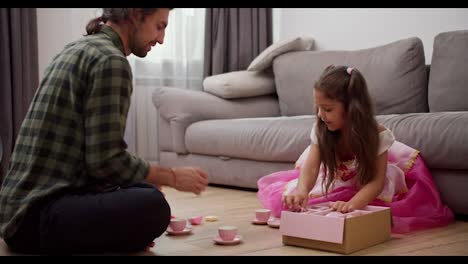 A-little-girl-in-a-fluffy-pink-dress-together-with-her-brunette-male-father-in-a-green-checkered-shirt-lay-out-toy-cups-for-tea-drinking-while-playing-at-home-in-a-modern-apartment