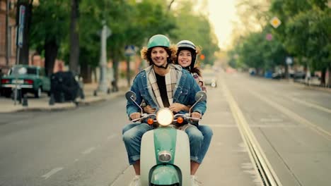 A-happy-guy-with-long-curly-hair-in-a-denim-jacket-rides-on-his-green-moped-with-the-headlights-on,-he-is-surprised-by-the-bumps-in-front-of-him-and-from-behind-the-guy’s-shoulder-his-happy-girlfriend-in-a-white-helmet-for-a-moped-looks-out-in-the-summer-on-a-wide-street-in-the-city