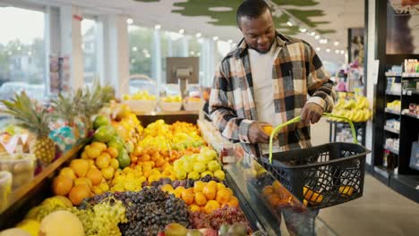 A-Black-skinned-brunette-man-in-a-checkered-shirt-and-a-white-T-shirt-chooses-citrus-fruits-and-tangerines-on-a-wide-counter-of-a-modern-grocery-store