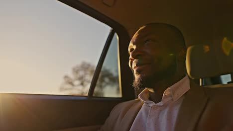 Happy-Black-skinned-businessman-in-a-brown-suit-returns-home-by-car-in-the-evening-after-a-hard-day-at-work-during-sunset.-A-man-with-Black-skin-is-driving-in-a-car-and-looking-out-the-window-at-the-flickering-rays-of-the-Sun-and-sunset