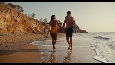 A-happy-couple,-a-blond-guy-and-his-blonde-girlfriend-in-an-orange-swimsuit,-walk-and-hold-hands-along-the-rocky-sandy-shore-of-the-sea,-they-hold-surfboards-in-their-hands-and-walk-along-small-waves-on-the-sea-morning-at-Sunrise