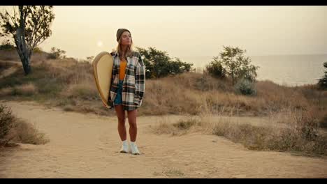 A-blonde-girl-in-a-plaid-shirt-stands-on-a-slope-near-the-sea-and-holds-a-wooden-surfboard-in-her-hands-against-of-the-sunrise-in-the-morning.-A-girl-stands-on-a-slope-near-the-sea,-inspects-the-place-and-waits-for-someone-to-surf-in-the-sea