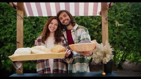 A-happy-brunette-guy-and-his-brunette-girlfriend-in-checkered-shirts-hold-out-trays-with-delicious-pastries-while-working-in-a-shop-against-the-backdrop-of-green-coniferous-trees-in-the-summer