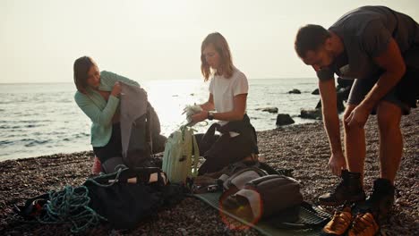 Two-blonde-girls-and-a-brunette-guy-in-a-gray-T-shirt-are-dismantling-their-backpacks-and-preparing-ammunition-to-create-a-belay-while-starting-their-rock-climbing-activity-on-a-rocky-seashore-in-summer.-A-group-of-rock-climbers-disassemble-their-backpacks-and-prepare-equipment-for-climbing-the-rock