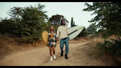 A-guy-in-a-hat-and-sunglasses-in-a-white-sweatshirt-walks-with-his-blonde-girlfriend-in-a-plaid-shirt-holding-hands-and-they-carry-surfboards-along-the-shore-near-the-sea.-Guy-and-girl-Surfers-go-down-the-slope-to-ride-and-swim-in-the-sea