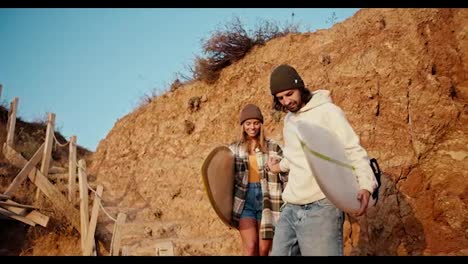 A-brunette-man-in-a-white-sweatshirt-helps-his-blonde-girlfriend-in-a-checkered-shirt-go-down-the-stairs-near-the-rocky-seashore.-They-carry-surfboards-in-their-hands-and-walk-to-the-sea-in-the-morning-at-Sunrise