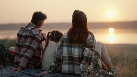 Vista-Trasera-De-Una-Familia-Feliz,-Marido,-Mujer-Y-Su-Pequeña-Hija-Sentados-En-Una-Colchoneta-Durante-Sus-Vacaciones-Y-Picnic-Fuera-De-La-Ciudad-Y-Mirando-La-Puesta-De-Sol-Cerca-De-Un-Estanque-En-La-Tarde-De-Verano.