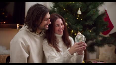 A-brunette-girl-in-a-White-sweater-shakes-a-New-Year's-glass-ball-together-with-her-brunette-boyfriend-near-a-green-New-Year's-tree-decorated-with-toys-and-a-garland-in-a-cozy-room-in-winter