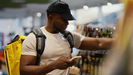 A-Black-skinned-food-delivery-man-with-a-large-yellow-bag-selects-the-necessary-goods-from-among-the-products-in-a-grocery-store-window.-A-delivery-man-checks-the-necessary-items-and-buys-them-to-deliver-with-a-large-yellow-bag