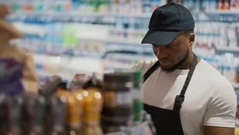 Close-up-shot-of-a-man-with-Black-skin-in-a-white-T-shirt-and-a-black-apron-laying-out-a-product-in-the-window-of-a-large-supermarket.-A-male-worker-at-a-grocery-supermarket-lays-out-goods-on-the-counter-and-sorts-them