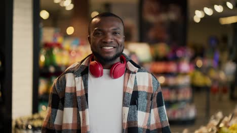 Portrait-of-a-happy-young-man-with-Black-skin-color-in-a-plaid-shirt-and-red-wireless-headphones-who-is-smiling-and-posing-near-the-counters-in-a-modern-grocery-store