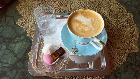 Small-tray-with-cappuccino-in-a-pastel-blue-polka-dot-cup,-some-sweets-and-a-glass-of-water-on-a-golden-table-coaster
