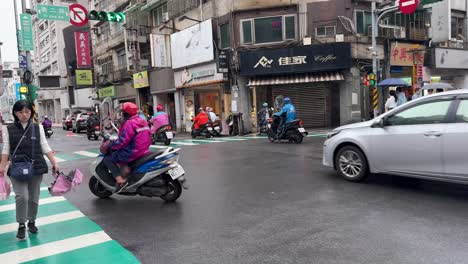 Panning-view-shot-of-Motorcyclists-wearing-raincoats-and-riding-on-a-rainy-day-and-pedestrians-crossing-in-Keelung-City,-Ren’ai-District,-Taiwan