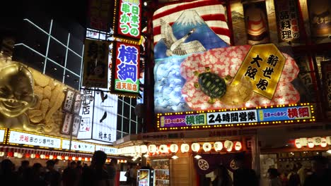 Disparo-Panorámico-A-La-Derecha-A-Través-De-Una-Colorida-Exhibición-Iluminada-Sobre-Un-Restaurante-Con-Una-Estatua-De-Biliken-En-La-Esquina-Del-área-De-Shinsekai-Por-La-Noche