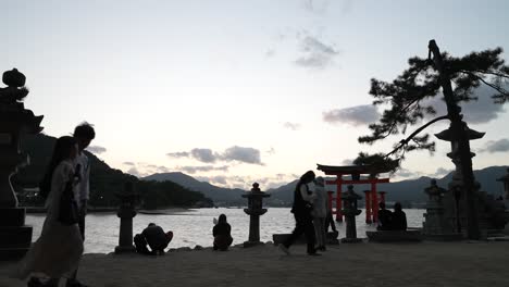 Touristen,-Die-Kurz-Nach-Sonnenuntergang-An-Miyajima-Vorbeigehen,-Mit-Jinja-Otorii-Im-Hintergrund-Hinter-Einem-Baum