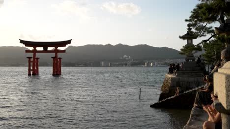 Goldene-Stunde-über-Dem-Torii-Tor-Von-Itsukushima-Mit-Touristen,-Die-An-Der-Wand-Sitzen-Und-Alles-In-Sich-Aufnehmen