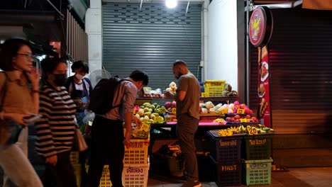 Two-people-busy-choosing-the-right-kind-of-fruits-to-buy-at-a-a-sidewalk-in-Bangkok-while-commuters-pass-by,-Bangkok,-Thailand