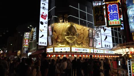 Night-Time-View-Of-Crowded-Busy-Street-Outside-Illuminated-Colourful-Display-Above-Restaurant-With-Biliken-Statue-On-Corner-In-Shinsekai-Area-At-Night