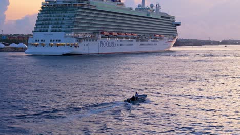 Skiff-drives-out-to-sea-alongside-large-cruise-ship-docked-at-port-in-Caribbean-ocean-at-dusk