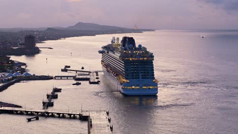 Rear-aerial-trucking-pan-of-cruise-ship-leaving-Caribbean-port-town-at-blue-hour