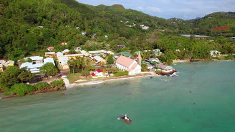 Drone-of-St-Joseph-church-near-the-shore-of-Anse-royale-beach,-cross-on-rock-in-the-center-of-the-ocean,-Mahe,-Seychelles-30fps-1
