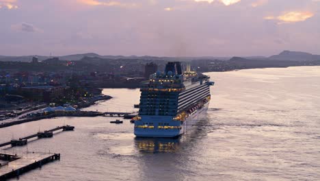 Rear-view-orbiting-around-cruise-ship-departing-from-port-town-in-Caribbean,-illuminated-at-epic-sunrise